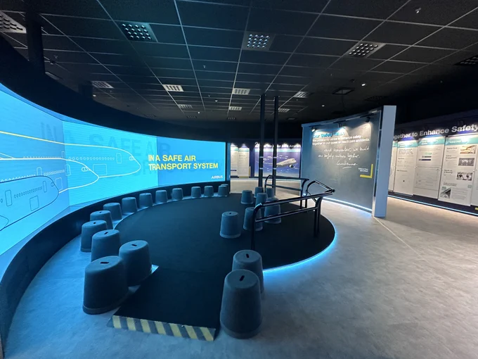 A large screen wraps around a room with seating. The screen reads: In a safe air transport system