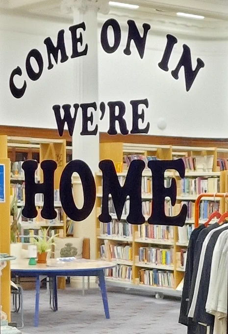 vinyl lettering on a window reads "Come on In We're Home", through the glass we see the main library space of the Glasgow Women's Library filled with stacks of books