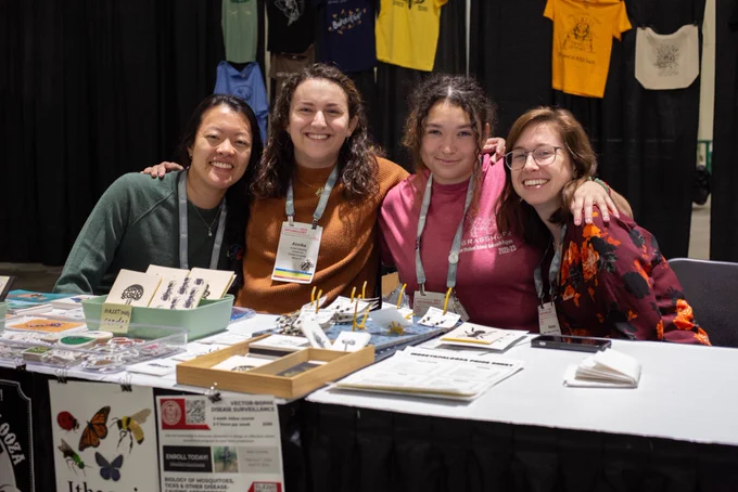 Four graduate students sitting behind a table with handmade insect-themes art and jewelry.