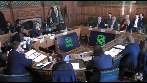 Group of people sitting around table with House of Commons logo. Small audience sitting behind them watching proceedings.
