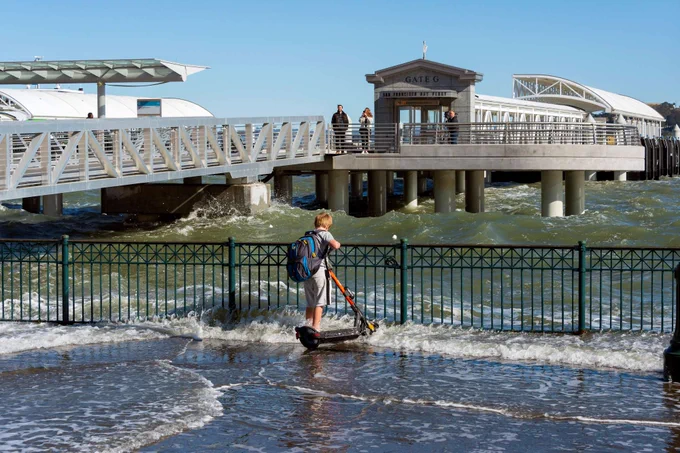 A boy stands with a scooter and looks over a railing at the Bay, which is overflowing below the railing onto the concrete where the boy stands. In the background is the pier for Ferry Gate G. It is a sunny day.