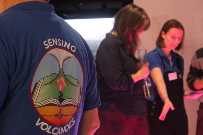 foreground: back of a man in a blue polo shirt with a logo on it. The logo has a volcano erupting out of a book, the centre of the book has a black crack down the centre and an off kilter house in front of the volcano. beneath the book is orange and red shades in which a seismic trace has been drawn and around the logo the words "sensing volcanoes" are written in all caps. In the background two women stand looking down, they are lit from below by a pinkish light and one woman is wearing a blue polo shirt and holding an ipad and gesturing downwards as if explaining something to the second woman. 