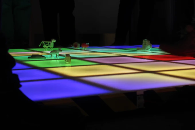 ground level view of a light up grid, shades of blue, yellow, pink green and red can be seen lighting up squares in a pattern. on some of the light up squares wooden toys can be seen dotted about the grid. Toys include a cow, a sheep, a camera, a pig, a set of binoculars, and a saw. 
