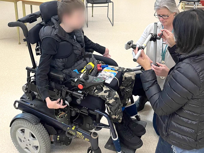 A student in wheelchair watches as two OT's attach and set up a mounting system for eye gaze to the wheelchair.