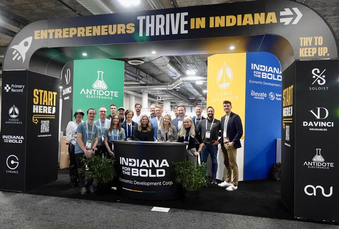A group of entrepreneurs and IEDC staff stands in front of the Indiana Economic Development Corporation's booth. "Indiana for the Bold" is on a stand in front of the group. An arch with the names of Indiana startups surrounds the group. "Entrepreneurs thrive in Indiana" is written on the top of the arch.