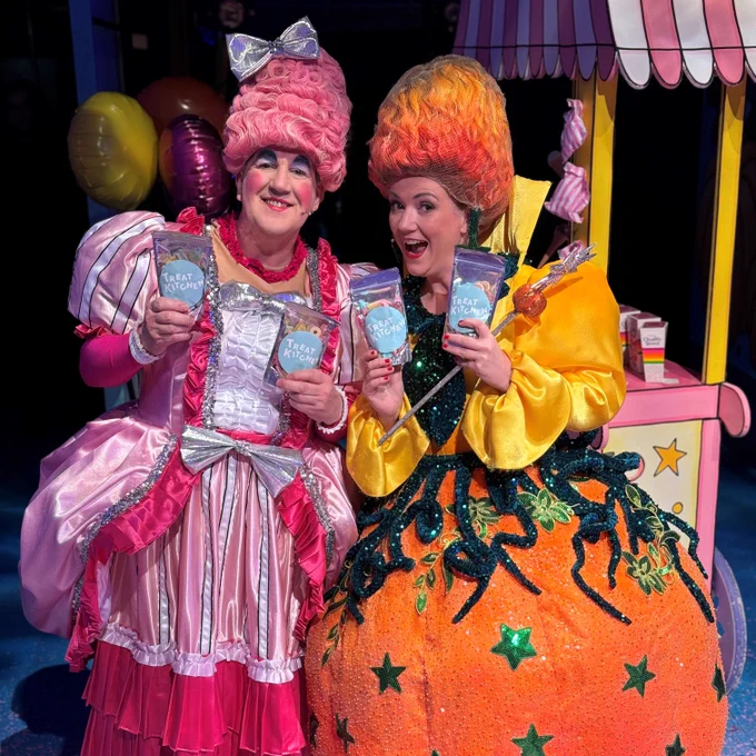 Pantomime Dame Rose in a pink dress and wig, and a Fairy Godmother dressed in orange with a starry orange pumpkin skirt and flame coloured wig holding packets of treat kitchen sweets and smiling . 