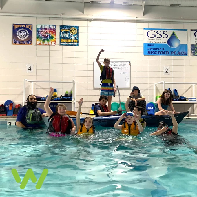 A group of nine middle school aged kids wearing life jackets and floating at the edge of a pool. One student in a kayak, all smiling and throwing their hands in the air.