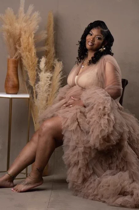 A pregnant Black woman smiles, wears a flowing light brown dress, sits in a chair and places her hands on her stomach. Her medium-long black hair is curled and falls over her shoulders. 