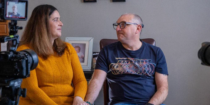 A man and woman sit on chairs, looking at each other. They are holding hands. A camera is in front of them, filming them.