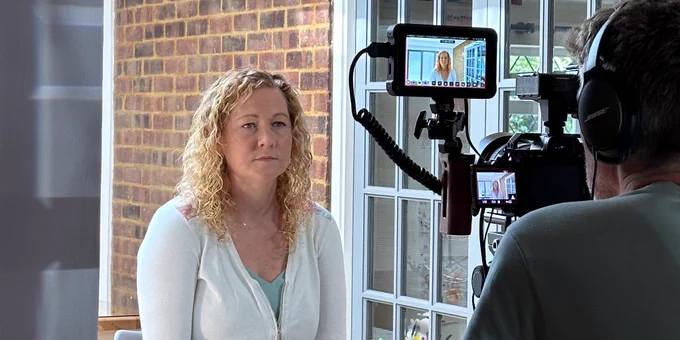 A woman sits. A camera is in front of her, filming her. A man wearing headphones looks into the camera.