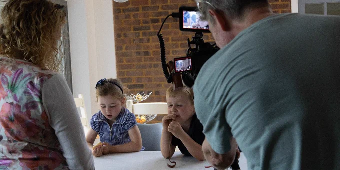 Two children sit at a table, eating. There is a camera in front of them. A man looks at the camera and a woman stands next to the table.