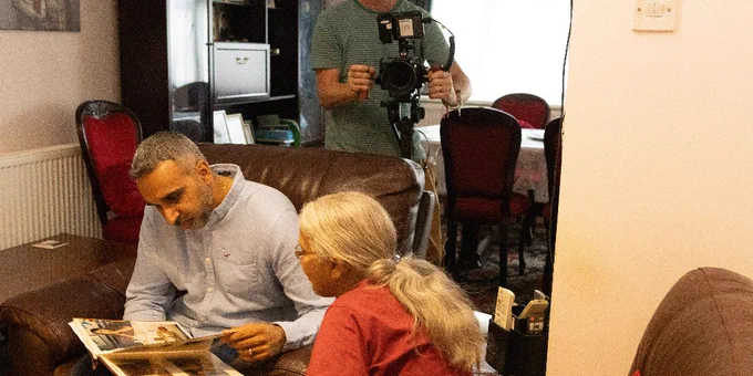 A man and older woman sit on armchairs, looking through a photo album. A man with a camera stands behind the armchairs, filming them.