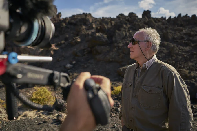 Werner Herzog stood in sunglasses in a rocky landscape, being filmed by a camera out of focus in the left hand corner of the frame.