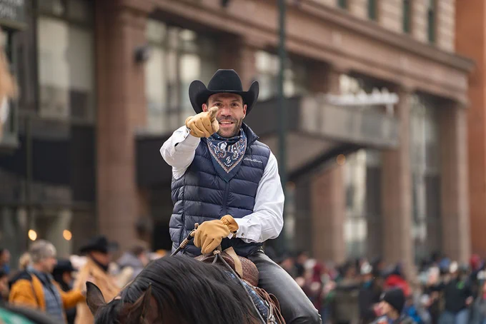 A person on a horse wearing a black hat, blue vest, bandana and gloves pointing at the camera while riding a horse in the National Western Stock Show kick off parade.