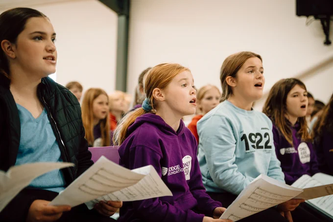 Only Kids Aloud members are seated in a rehearsal at their residential in Llangrannog.  They are holding sheet music.