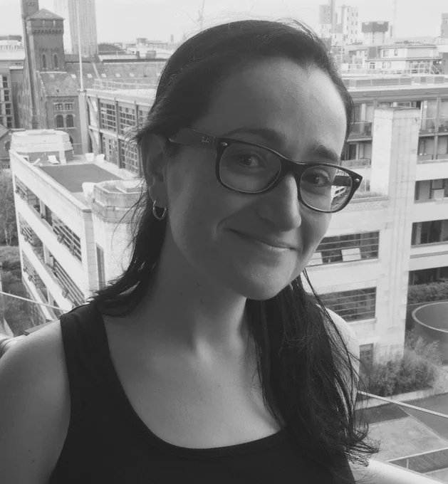 Black and white photo headshot of a woman with long dark hair and glasses. She is smiling at the camera and there is a skyline of a city.