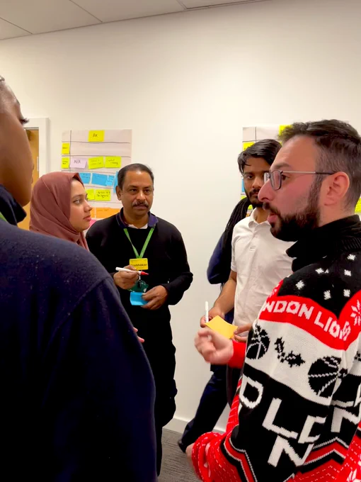 A group of people standing in a circle talking. They are holding post it notes and pens. Behind them are swim lane process maps using flip charts and post it notes