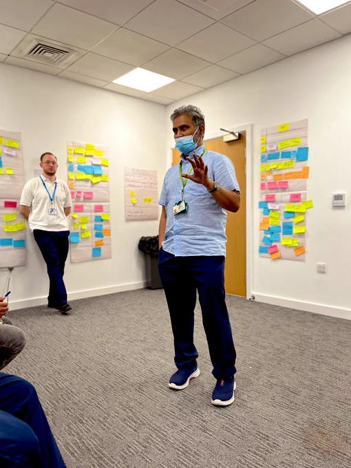 A man wearing a mask and speaking to a room of people. Behind him are swim lane process maps using flip chart and post it notes