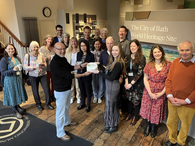 A group of staff and volunteers in the Bath World Heritage Centre. Two staff members are standing in the middle of the image holding a cake with the Bath World Heritage logo on it.