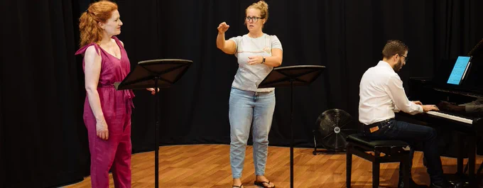 A woman in a burgendy jumpsuit stands in front of a music stand. She is looking at another woman who is using their hands to sign in British Sign Language. A man is also sat at a grand piano playing. 