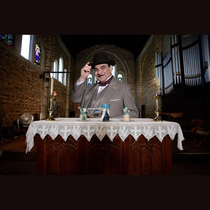 Image is of a church with the altar at the front. Behind the altar instead of the usual priest is the actor, David Suchet portraying the character of Hercule Poirot.