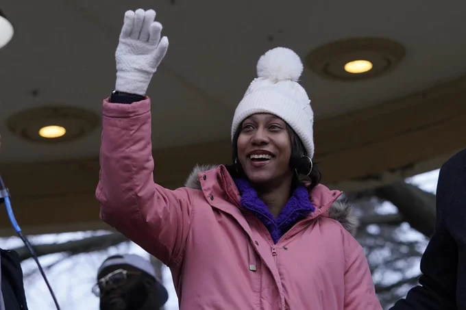 Brittany watts, smiling and wearing a pink coat, white hat and white gloves as she waves to the crowd