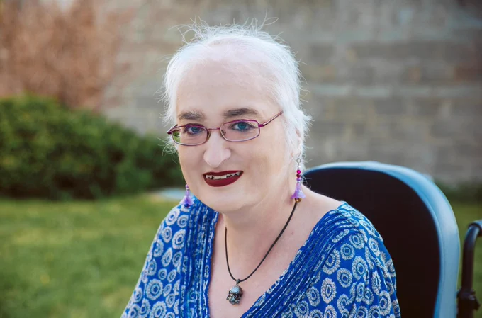 Beautiful lady with short white hair, dark eyebrows, rich red lipstick, pink metallic glasses, sitting in a chair, with blue white flower patterned v-neck top, trendy pink red drop earrings and non-matching  multicoloured stone on a leather necklace. With blue twinkling eyes and warm smile on her young unlined face.