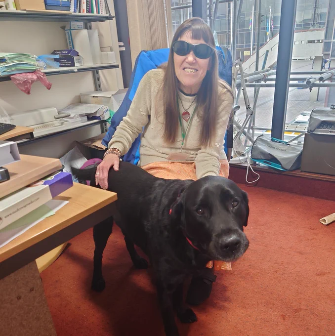 Photo of Visual Impairment Officer at Portsmouth Libraries, Julie Duffy, sat on a chair wearing dark glasses and smiling at the camera. She has her hand on a black Labrador who stands in front of her.
