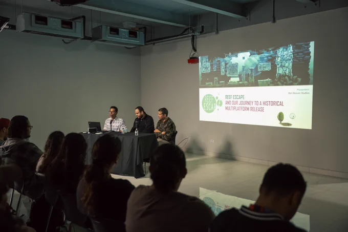 Fotografía de el ex alumno Jonathan Martínez, el profesor José Ramos y el profesor Miguel Santiago, sentados en una mesa hablando con una proyección atrás que lee "Reef Escape: And our journey to a historical multiplatform release".