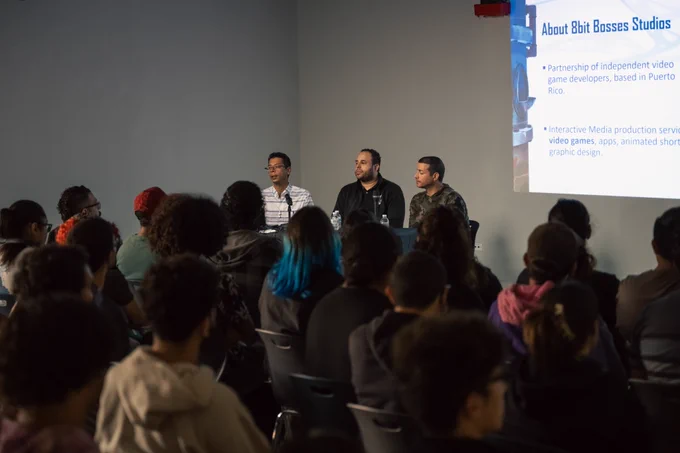 Fotografía de el ex alumno Jonathan Martínez, el profesor José Ramos y el profesor Miguel Santiago sentados hablando con el público con una proyección atrás que lee "About 8bit Bosses Studios. Partnership of independent video game developers, based in Puerto Rico. Interactive Media production services, video games, apps, animated shorts, graphic design".