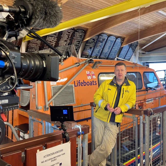 A Selsey RNLI crew member wearing a yellow waterproof jacket and beige trousers standing in front of an orange lifeboat, being filmed in the station with a black camera in the foreground.