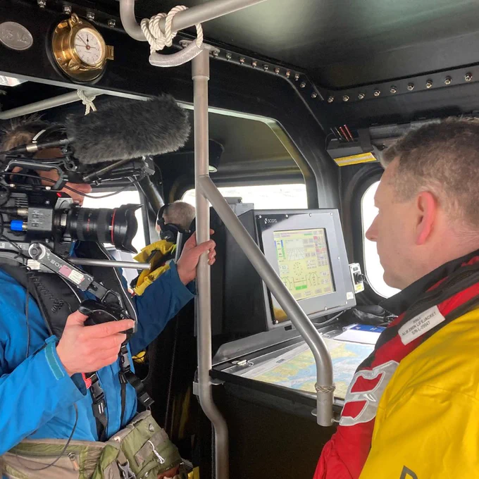 A cameraman wearing a blue jacket filming a Selsey crew member dressed in a black and yellow drysuit aboard an all weather lifeboat. Instruments and chart table are in the background.