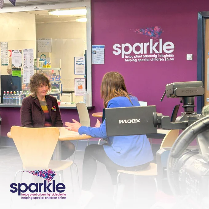 An image of our Founder Dr Sabine Maguire sat at a table opposite a reporter from ITV who has her back to the camera. They sit in front of a purple wall with the 'Sparkle' logo on, and in the fireground there is a camera pointing at them.