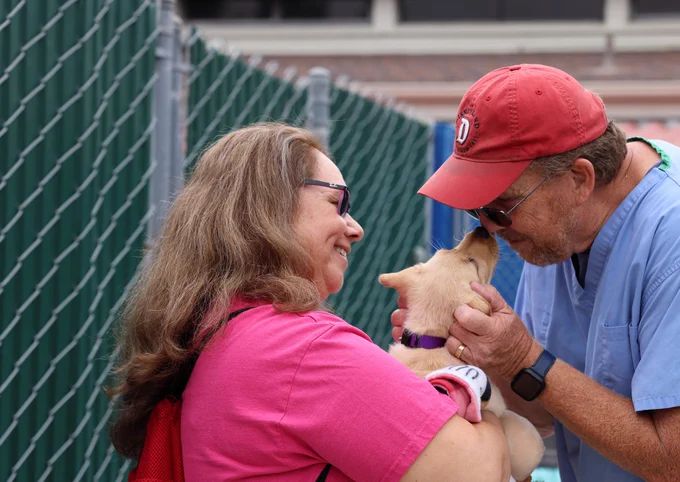In this photo, a woman holds a yellow Lab puppy and a man leans into the puppy for a kiss.