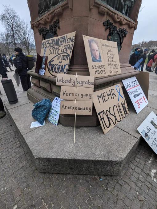 Papp-Plakate lehnen an einer Steinsäule. Auf ihnen steht zB: Lebendig begraben. Versorgung. Anerkennung. mehr Forschung. SEVERE MECFS