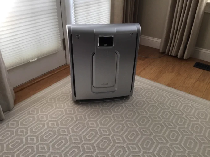 A BioGS Classic air purifier stands in a room with natural light filtering through blinds, complementing a neutral-toned rug and hardwood floors.