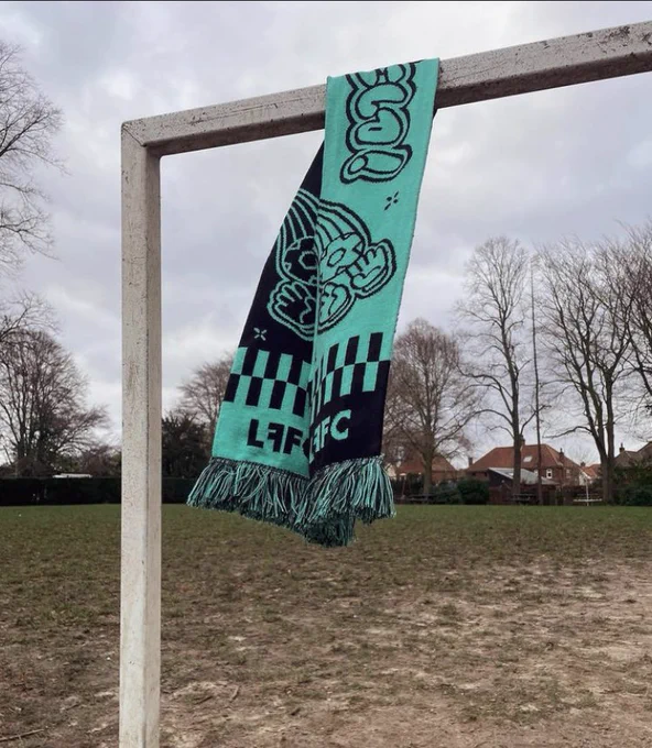 A teal and black patterned scarf, embroidered with the letters “LFFC”, hangs from the top bar of a net-less football goal. The scene is slightly dystopian: trees barren and dying against an overcast (nuclear cloud) sky while the once-grassy pitch below shows only remnants of green and resembles the moisture barren, cracked dirt swathes off the Mojave Desert. There are no humans in sight…
