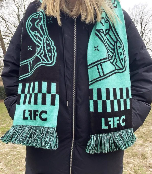 A headless and handless human in a thick black winter coat has a teal and black patterned scarf, embroidered with the letters “LFFC”, draped around their neck.
