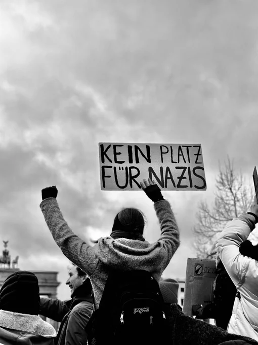 Foto einer Person mit Winterjacke und Rucksack von hinten. Sie reckt die linke Faust zum Himmel. In der rechten Hand hält sie ein Schild “Kein Platz für Nazis”. Im Hintergrund sind Teile des Brandenburger Tors zu sehen.