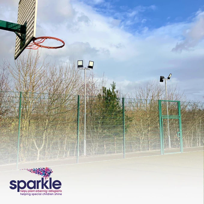 An image of the MUGA court. A basketball hoop is in the foreground in the top left corner and in the background you can see the lights overlooking the court.