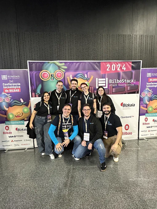 Parte del equipo de Lean Mind posa sonriente delante del cartel de bilbostack 2024 todas y todos van vestidos con las sudaderas  y polos de Lean Mind de color negro.En la foto están Ana, Adrián,Airan,María,Ari de pie y abajo agachados Miguel,Cristian y Manu.