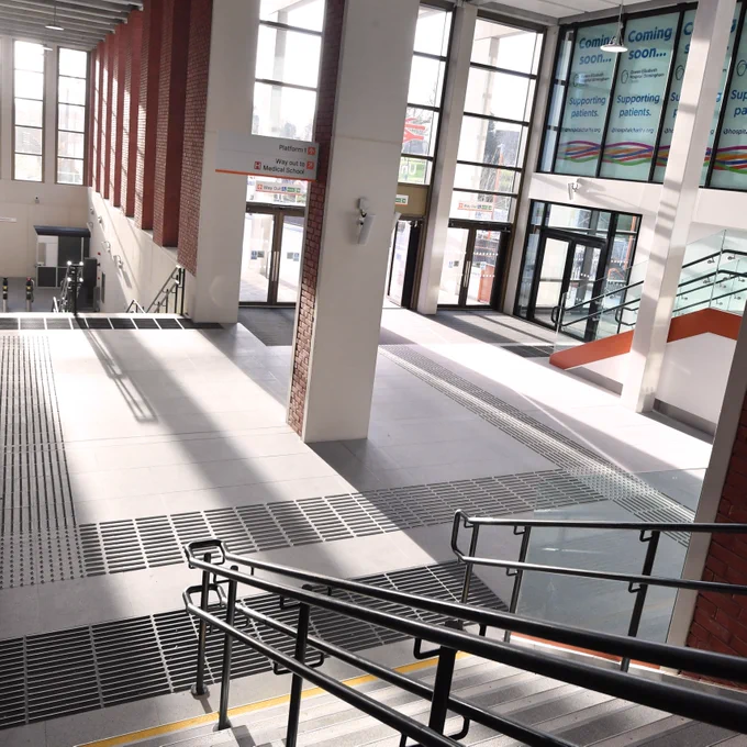 An image of the new University station building with mulitople extra side staircases and double bar handrails, double height ceiling above, large windows which flood the concourse with light and a multi door entrance / exit. 