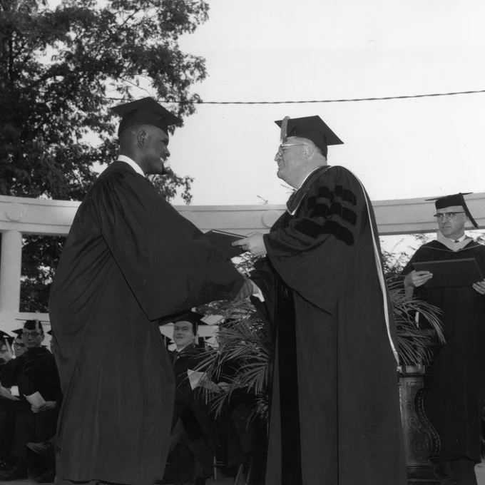 Harvey Gantt on the left shaking hands and receiving a diploma with people wearing graduation attire.