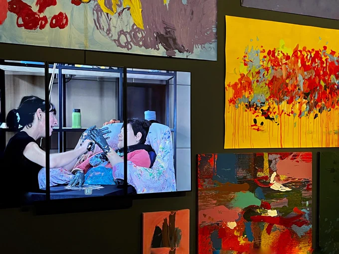 One small section of the extensive exhibition space on the ground floor of Baltic Gateshead. This shows one of the film screens situated on the wall with the art work. Picturing two people interacting through the creative process and both have hands liberally covered in dark paint. Between them is a part painted art work. The artist creating the work is sitting in supportive seating covered with a paint splattered sheet in a studio space. The word ‘Hi’ can just be seen at the base of the screen. To the right and above are colourful art works by Joe Goldman, Sam Smith, Gemma and Louise Newham.