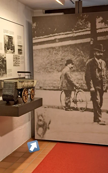 Ein großes, historisches Foto als Wandbild einer Ausstellung über die Entwicklung von Automobilen. In der untersten linken Ecke des Fotos rennt, unscharf zu sehen, ein Hund aus dem Bild. 