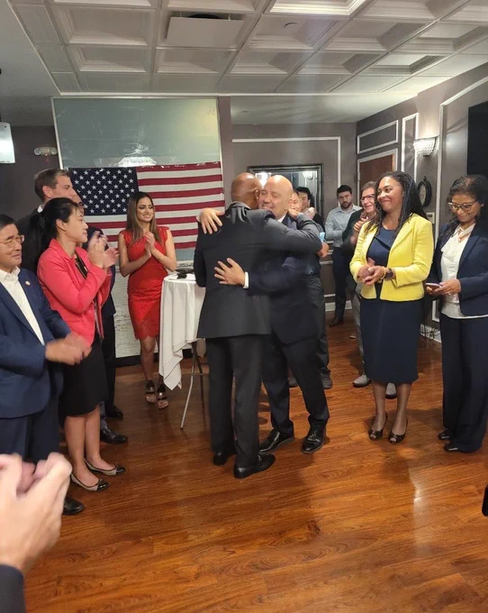 Mayor Adams hugs Paul Vallone. Many people around them applaud.