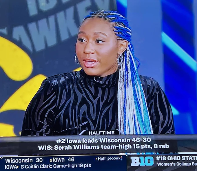 Photo of Aliyah Boston as a halftime analyst for Peacock. She has long blue and white braids, and she’s wearing a black dress that has tiger-print stripes, also in black.