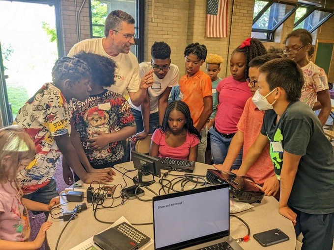 A group of children are gathered around a table with laptops.