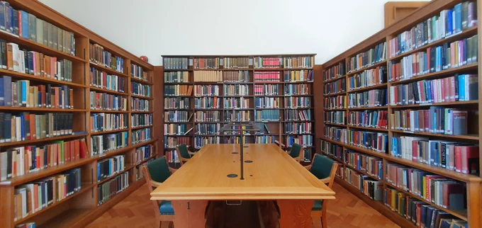 A large desk with a central light and four chairs upholstered in green leather sits in front of a large bookcase filled with books.