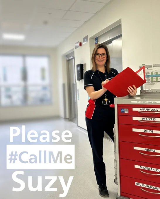 A friendly woman, holding a red folder, whilst looking at the camera. She is leaning against a red resuscitation trolley in hospital.
